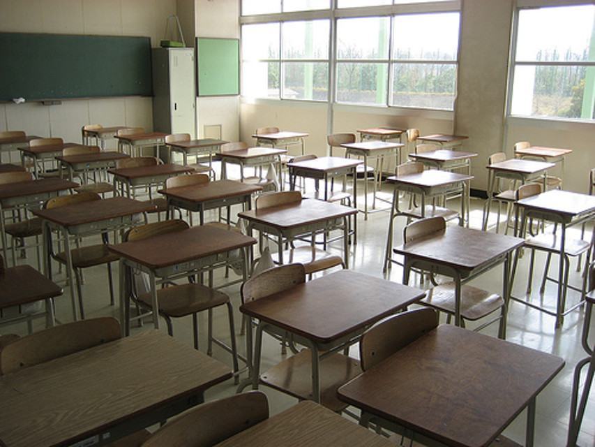 Aula scolastica vuota con banchi e sedie in legno davanti a una lavagna e grandi finestre