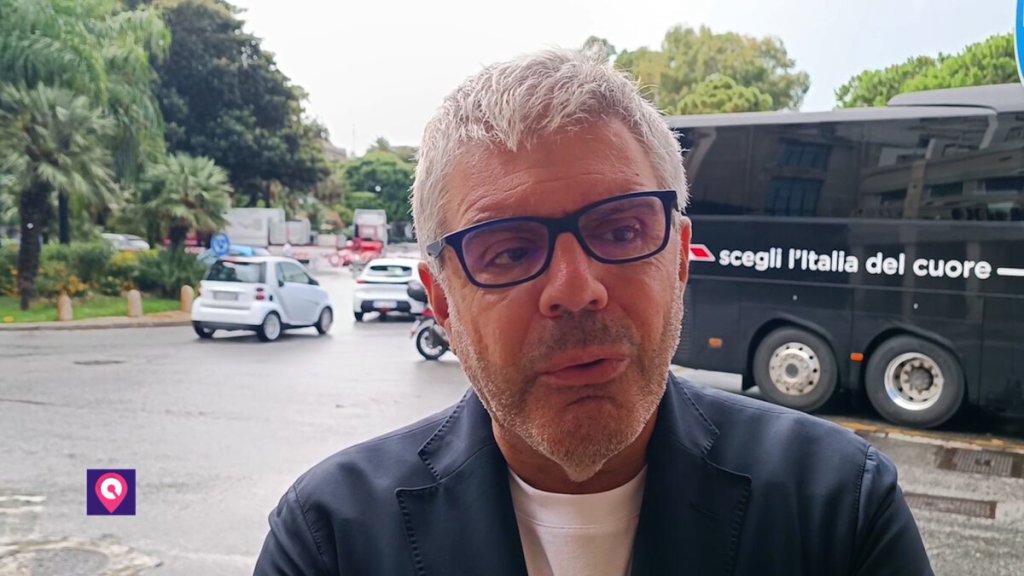 Il promoter Ruggero Pegna parla davanti alla telecamera in strada, con un autobus scuro sullo sfondo