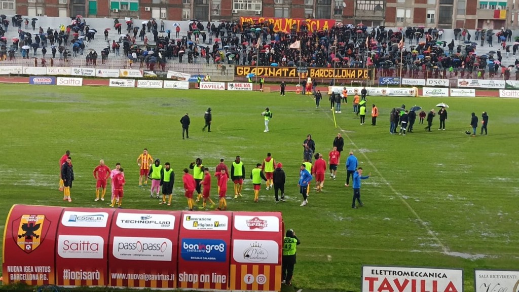 Campo allagato dello stadio D'Alcontres-Barone nel rinvio di Igea Virtus-Reggina, con tifosi sotto gli ombrelli
