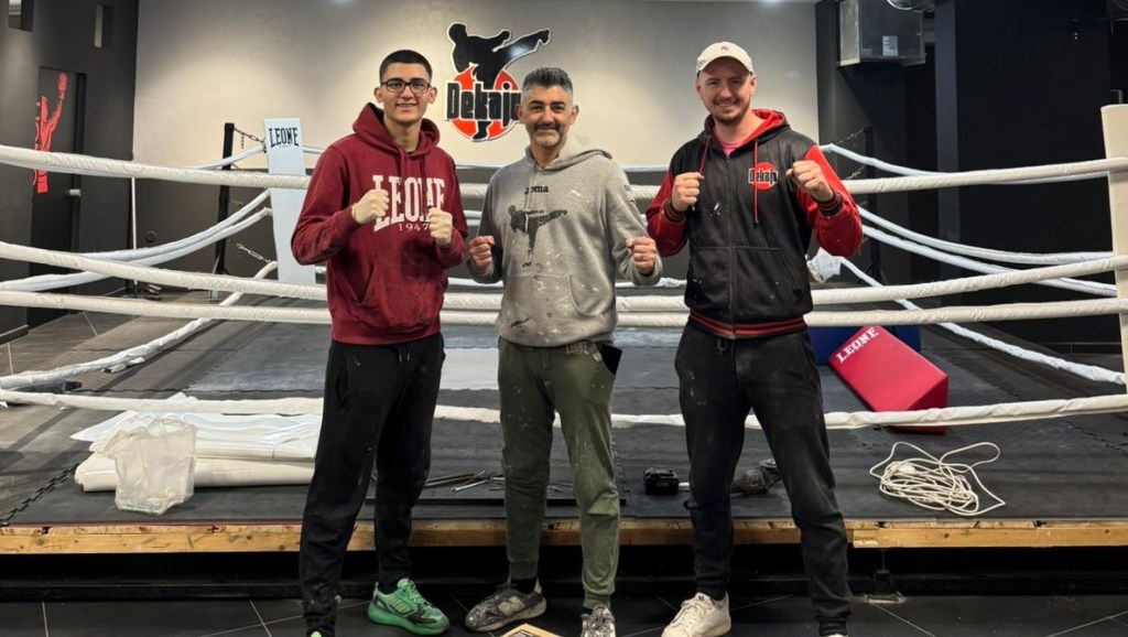 Tre uomini con i pugni alzati davanti al ring della palestra Dekaju Kombat, sotto il logo Dekaju