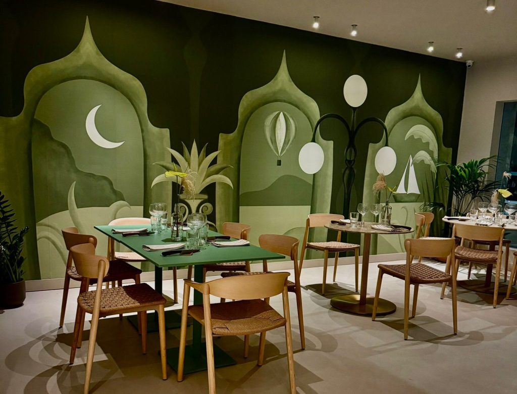 Sala di Mysia Bistrot con tavoli apparecchiati e un grande murale verde con luna, mongolfiera e barca a vela