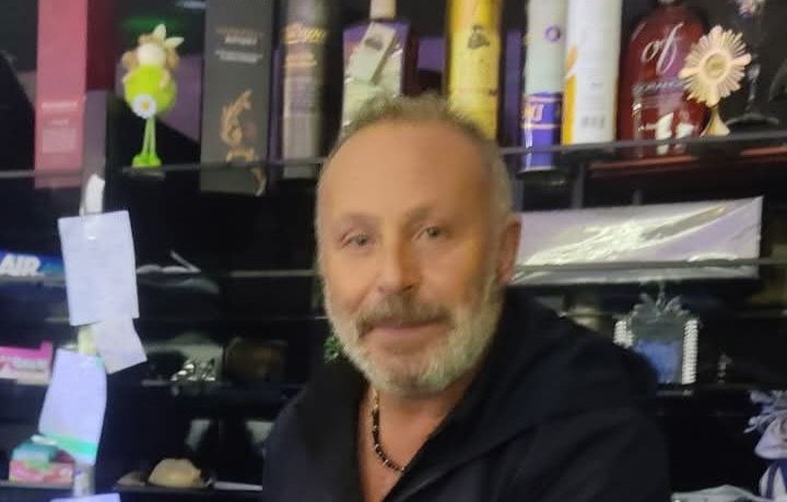 Uomo con barba grigia in un locale, davanti a scaffali con bottiglie e prodotti, nel bar Ninni's