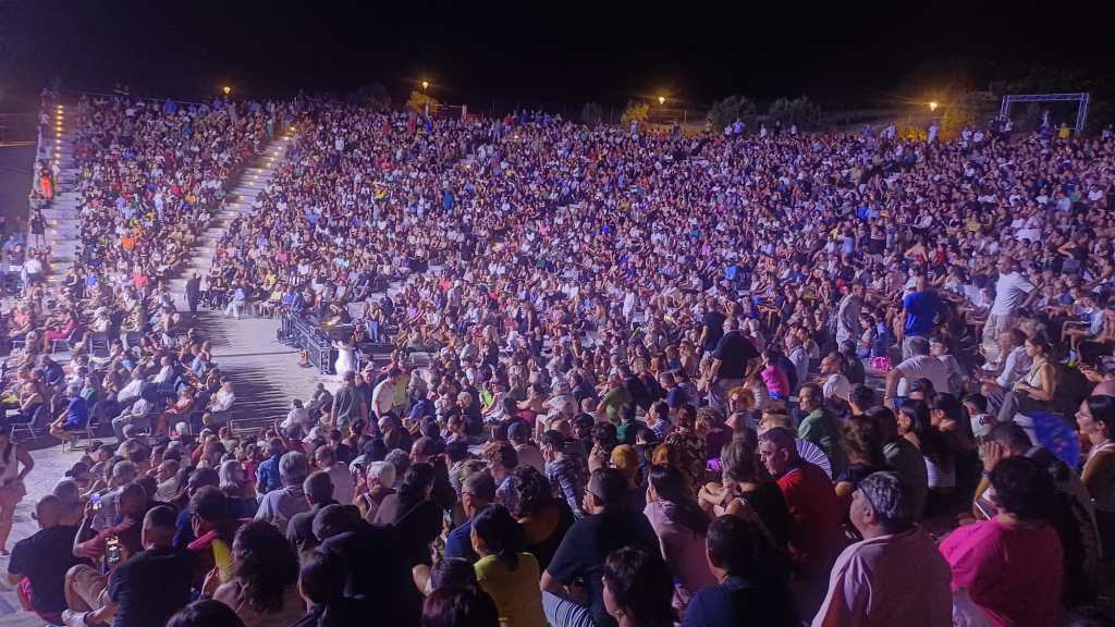 Il pubblico riempie le gradinate dell'Arena Epizefiri durante uno spettacolo serale, in una luce viola