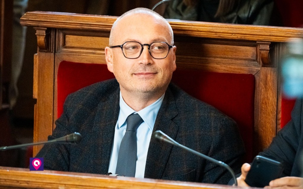 Uomo con occhiali, giacca scura e cravatta seduto su una panca di legno con schienale rosso, con due microfoni davanti