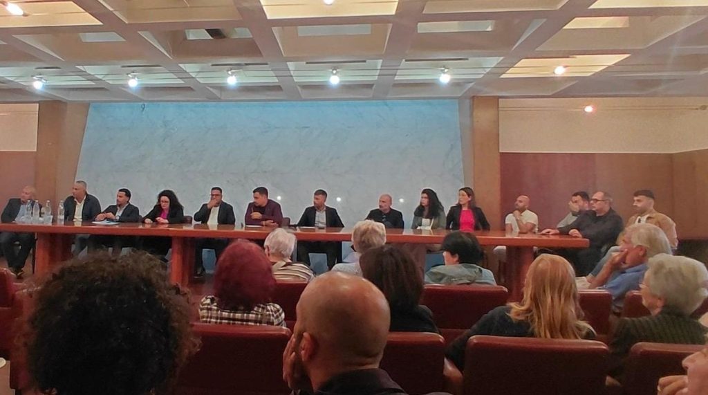 Assemblea pubblica in una sala: i relatori seduti a un lungo tavolo e il pubblico in platea in primo piano
