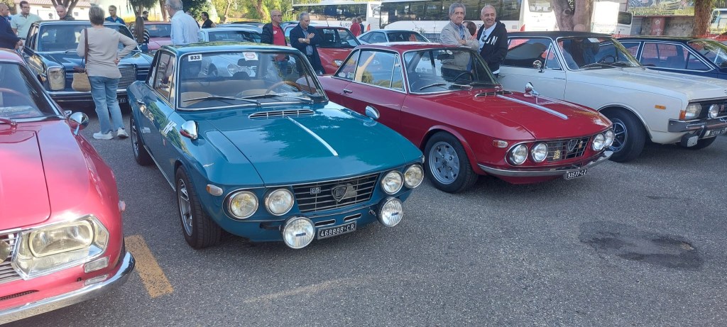 Auto sportive d'epoca parcheggiate affiancate, tra cui una coupé azzurra e una rossa, con alcune persone sullo sfondo