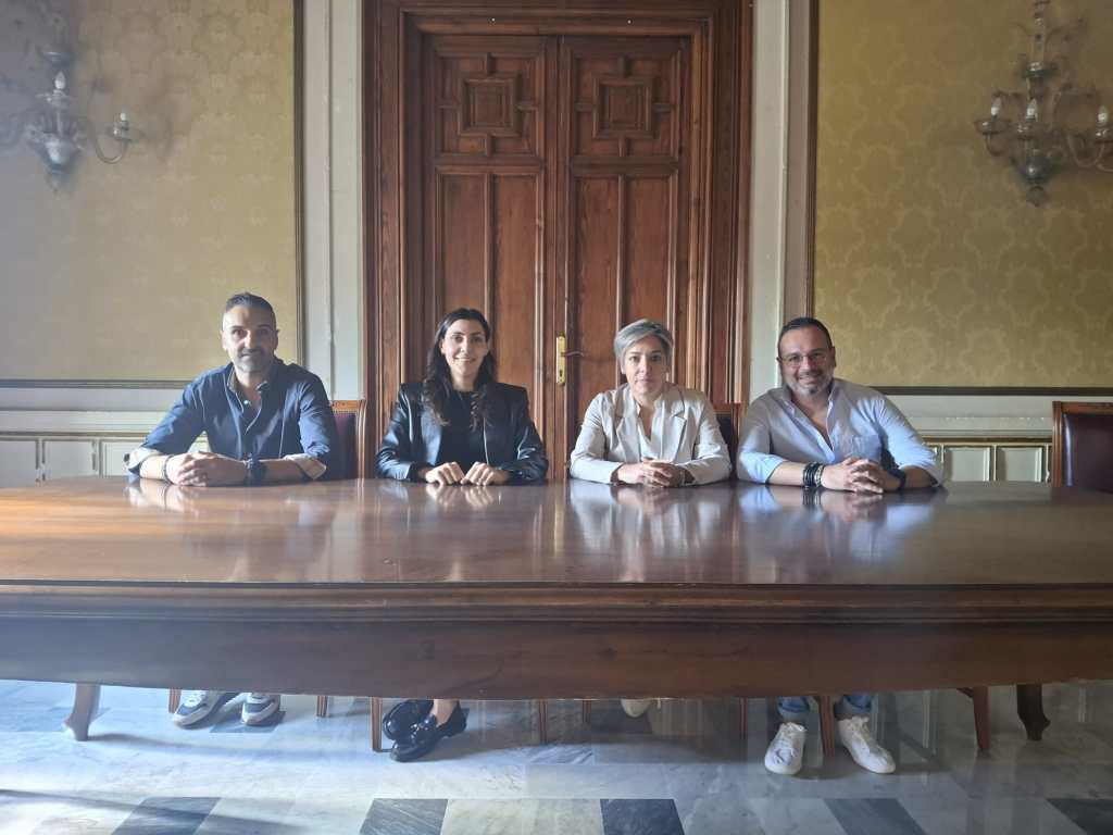 Quattro consiglieri seduti dietro un lungo tavolo in legno, in una sala con porta intagliata e lampadari alle pareti