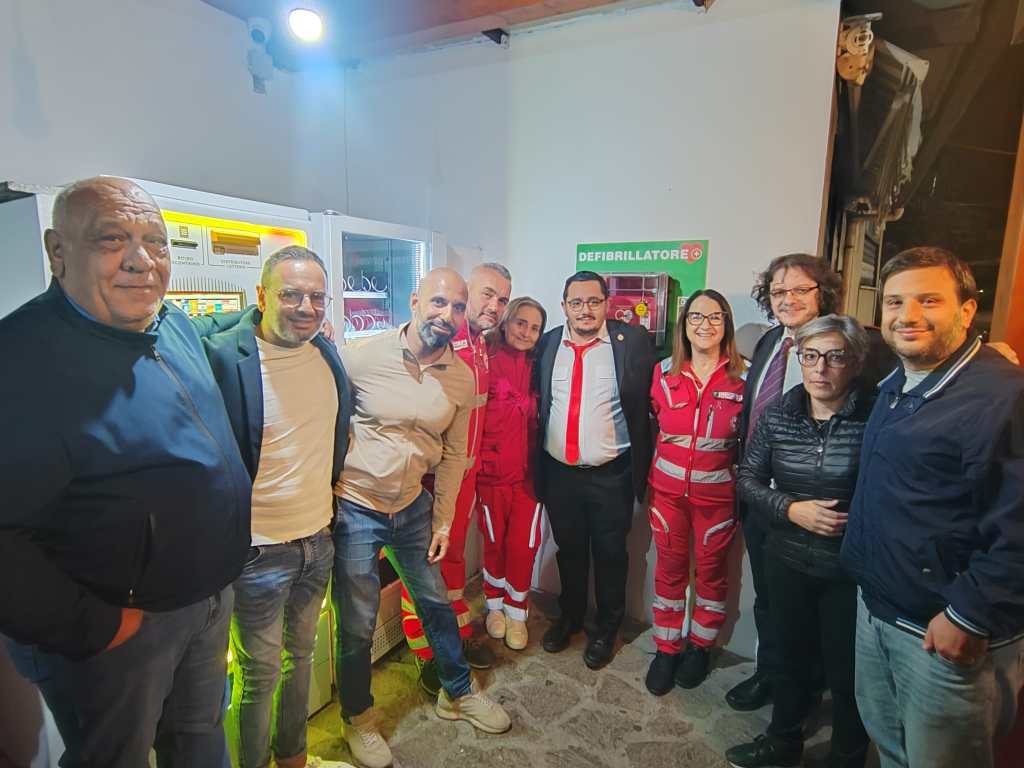 Persone e volontari in divisa rossa accanto al defibrillatore installato a parete, sotto il cartello verde Defibrillatore