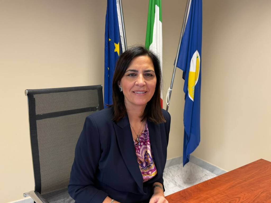 Una donna con giacca blu e camicetta a fantasia seduta a una scrivania, con alle spalle le bandiere dell'Unione europea, dell'Italia e della Calabria