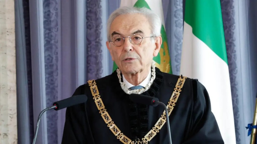 Un uomo anziano con occhiali, in toga nera con collare dorato, parla davanti a due microfoni con la bandiera italiana alle spalle