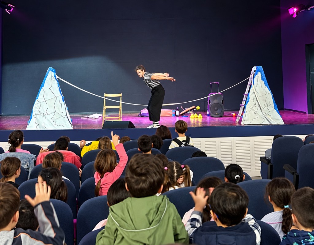 Un attore in maglia a righe sul palco tra due rocce di scena e una corda, davanti a una platea di bambini seduti in sala