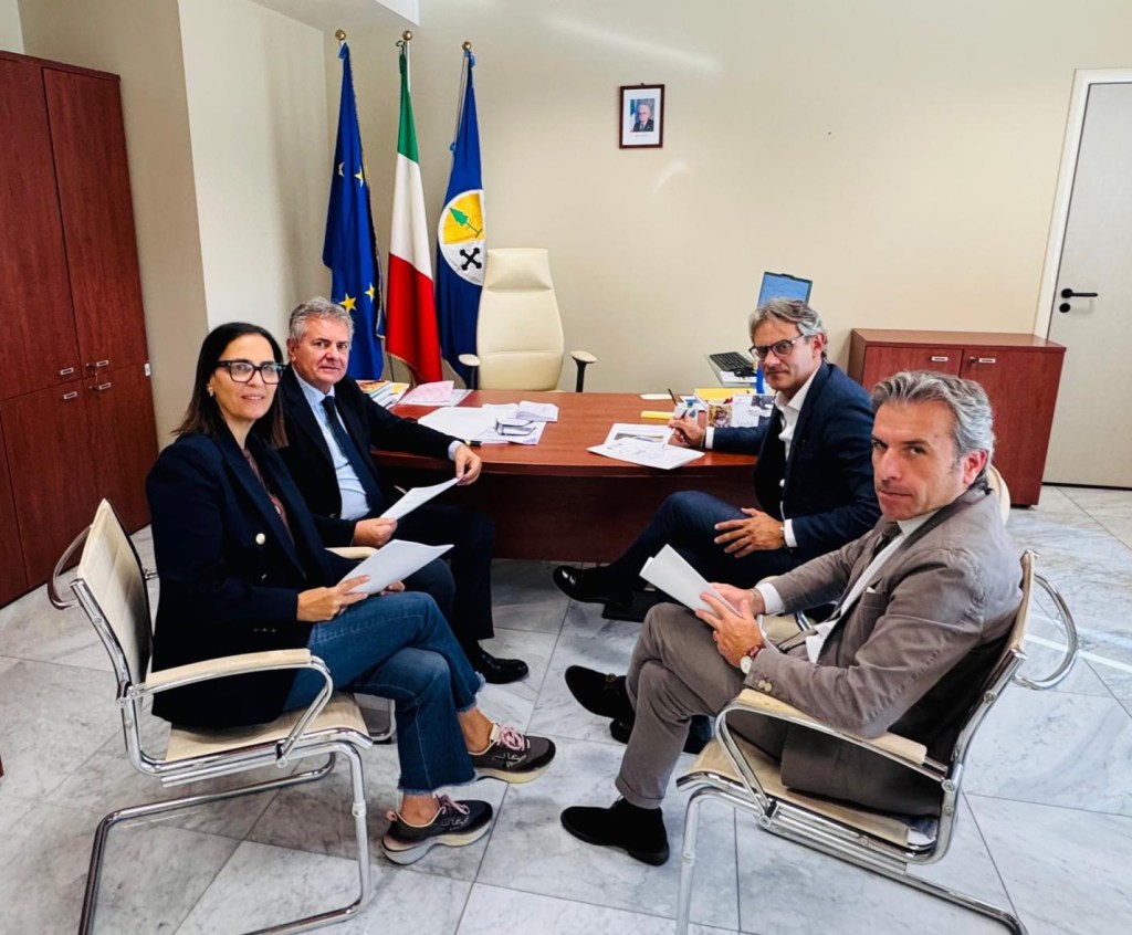 Quattro persone con dei fogli in mano attorno a una scrivania in un ufficio con le bandiere dell'Europa, dell'Italia e della Calabria