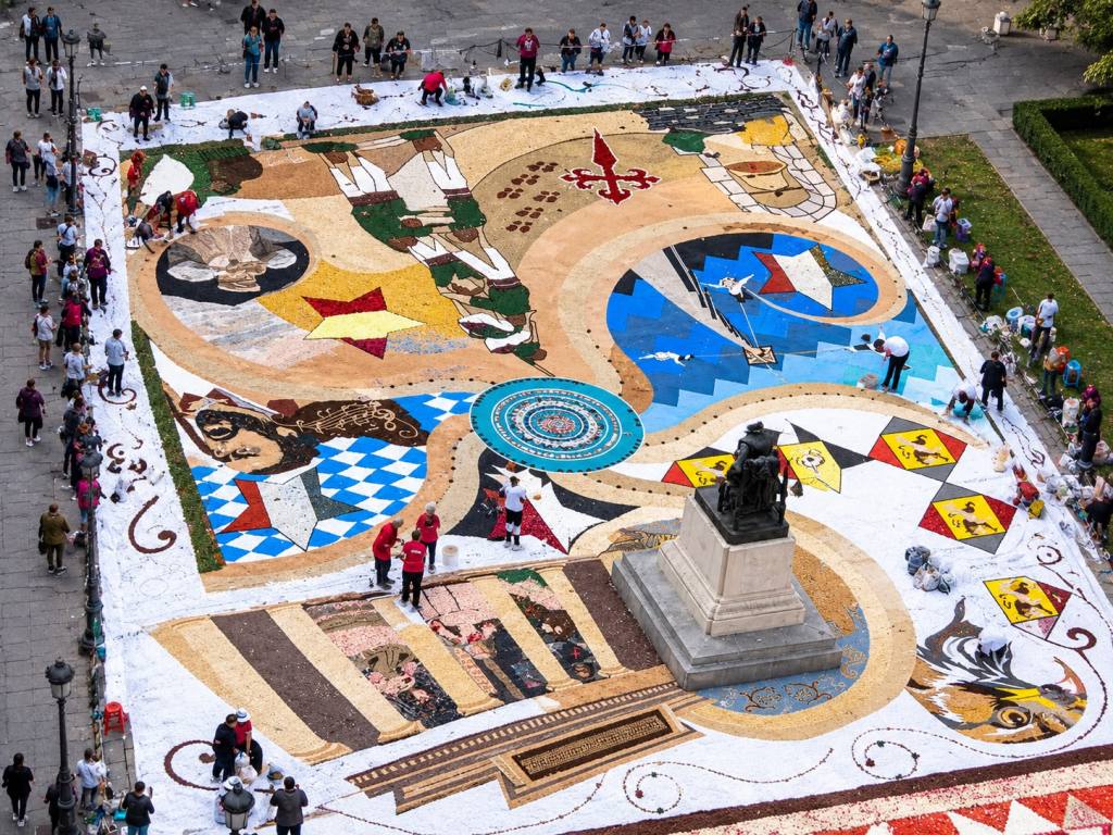 Grande tappeto floreale di arte effimera realizzato attorno a una statua, con numerosi infioratori al lavoro, visto dall’alto