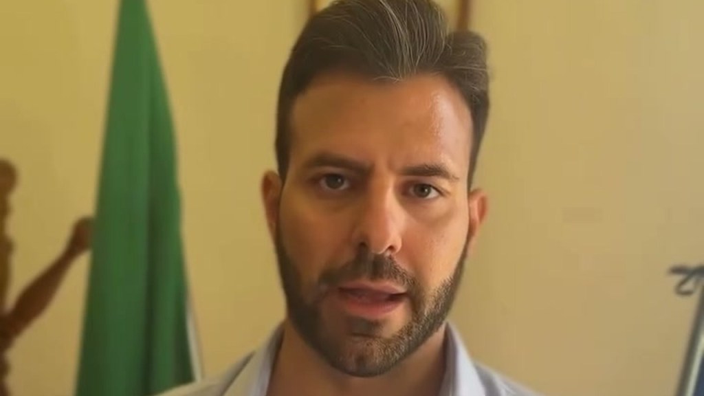 Un uomo con barba e camicia azzurra parla guardando in camera davanti a una bandiera verde