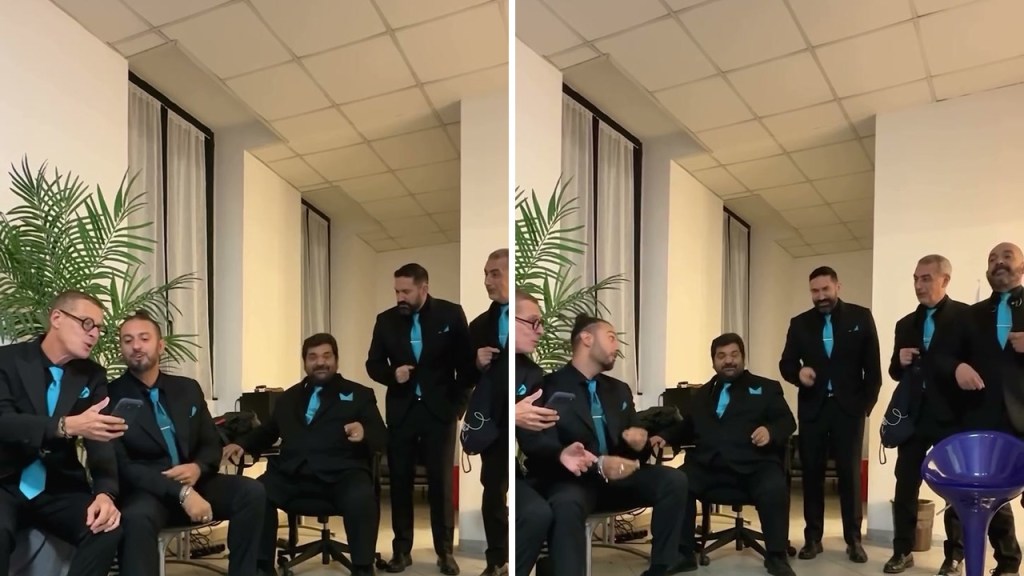 I Neri per caso in abito nero e cravatta azzurra in una sala, alcuni seduti e altri in piedi, durante l'esibizione a cappella
