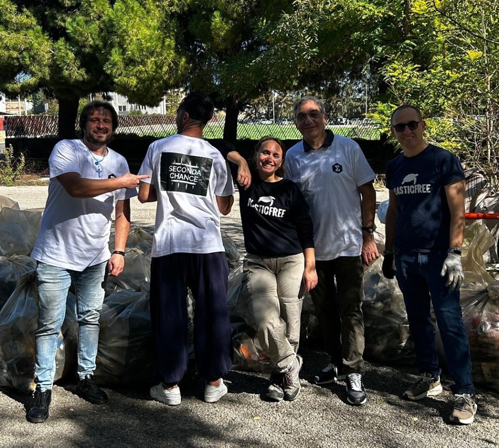 Cinque persone in posa con magliette Plastic Free e Seconda Chance davanti a sacchi pieni di rifiuti, sotto gli alberi