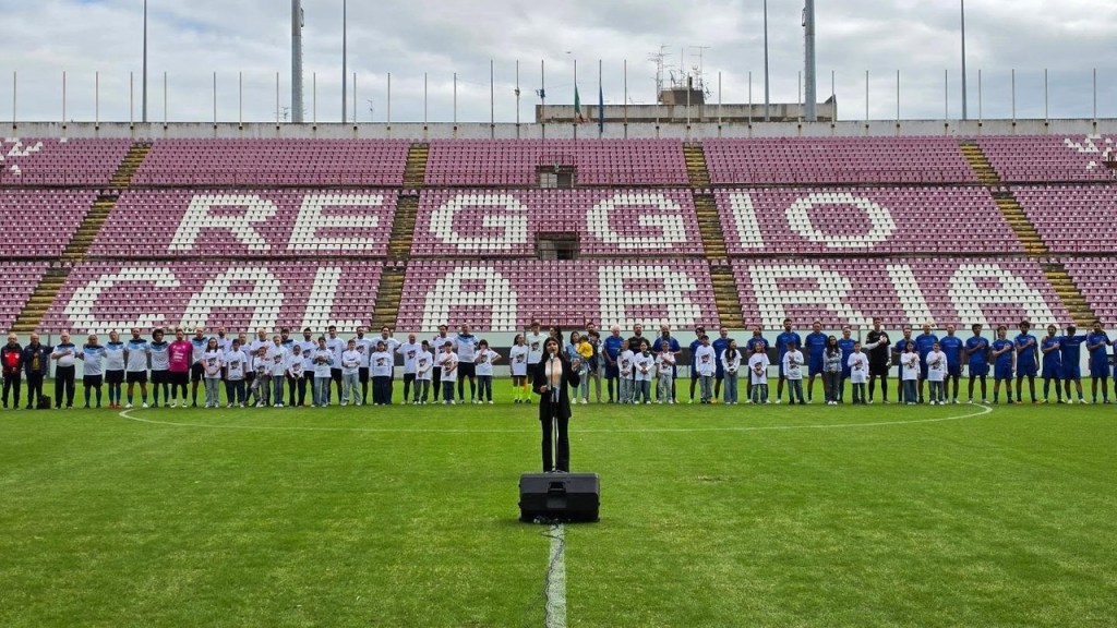 Giocatori e bambini schierati sul campo davanti alle tribune con la scritta Reggio Calabria