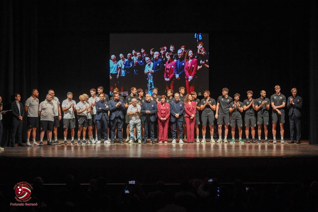 Atleti, staff e persone in abiti eleganti schierati in fila sul palco del teatro, con un maxischermo alle spalle