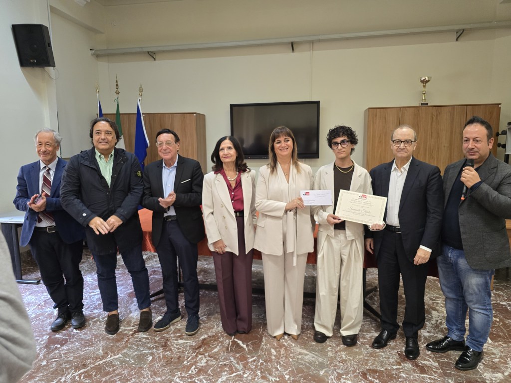 Un giovane con l'attestato della borsa di studio insieme a sette persone, in un'aula con bandiere e un trofeo sullo sfondo