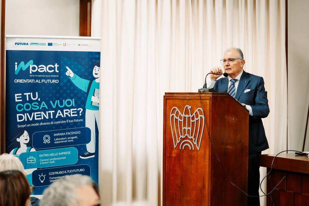 Un uomo in giacca blu parla al microfono dietro un leggio con l'aquila di Confindustria, accanto a un roll-up Impact Orientati al futuro