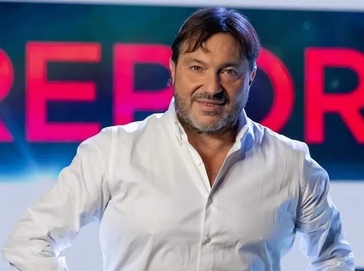 Sigfrido Ranucci in camicia bianca sorride davanti a uno schermo con la scritta Report in rosso