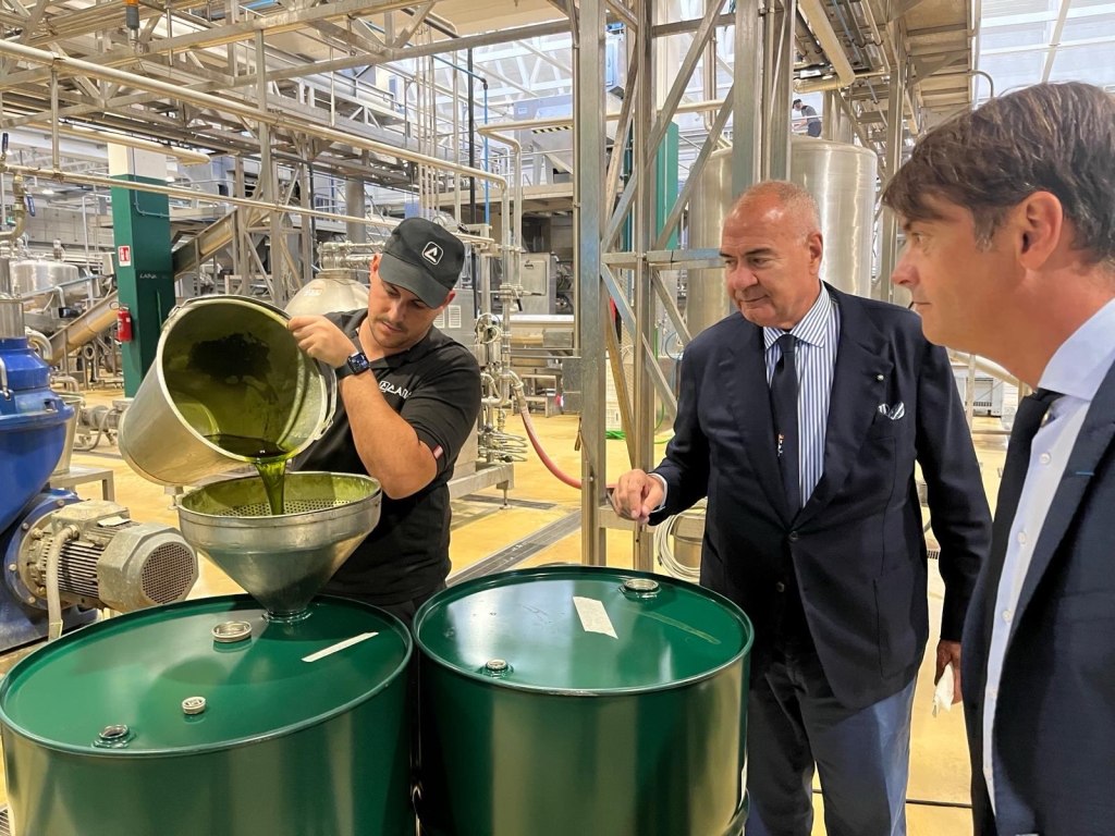 Un operaio versa olio essenziale verde in un imbuto su un fusto, osservato da due uomini in giacca nello stabilimento