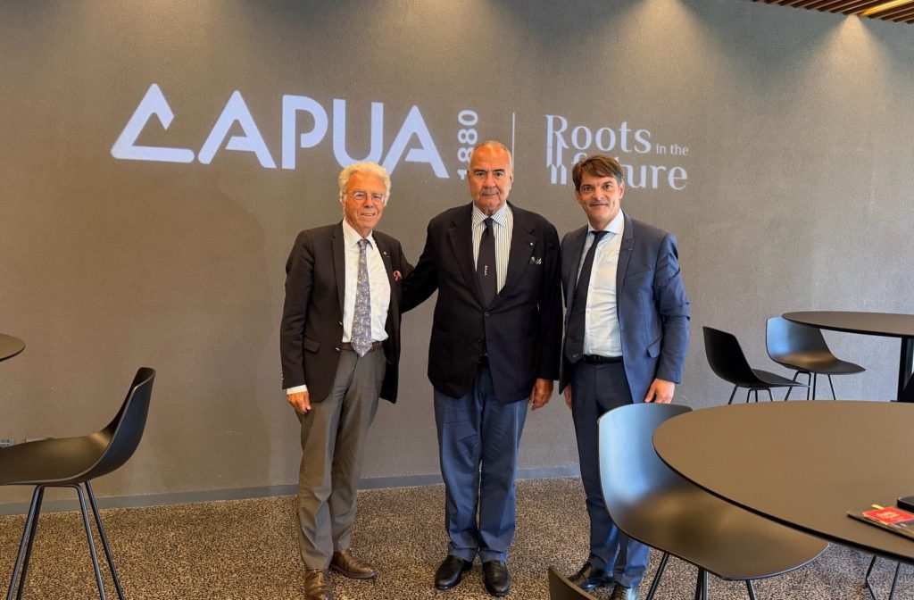 Tre uomini in giacca e cravatta posano davanti alla parete con il logo Capua 1880 e la scritta Roots in the future