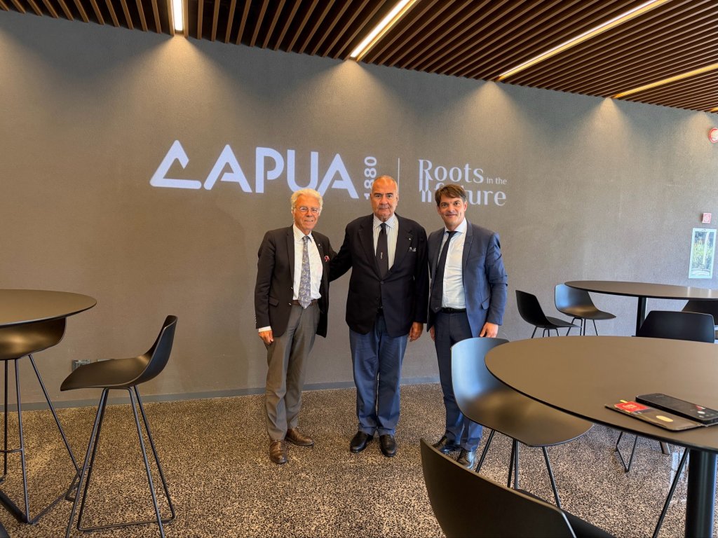Tre uomini in giacca e cravatta posano davanti alla parete con il logo Capua 1880 e la scritta Roots in the future
