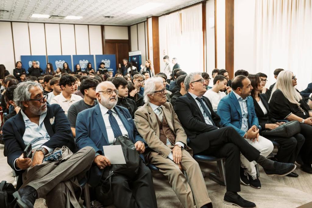 Sala affollata di studenti e ospiti seduti, con in fondo i pannelli blu con il logo di Confindustria
